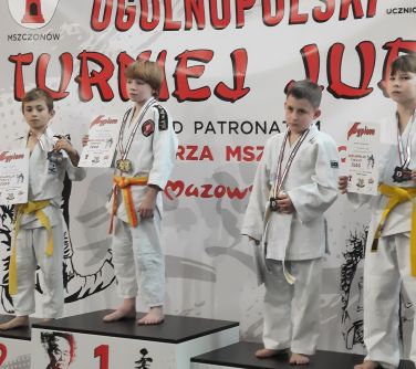 Hala. Czworo dzieci pozuje na podium. Dzieci ubrane w kimona do judo. W rękach trzymają dyplomy. Na szyjach medale