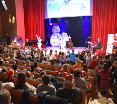 Aula. Publiczność siedzi. Na scenie osoby występujące. W tle dekoracja świąteczna