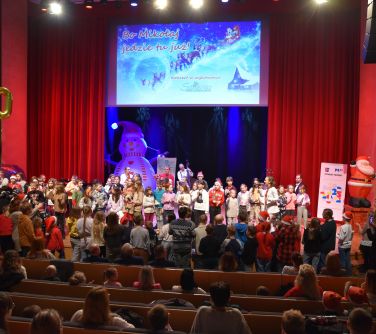 Aula. Publiczność siedzi. Na scenie występujące dzieci. W tle dekoracja świąteczna