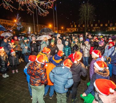 Na dworze wieczór. Dzieci w czapakach mikołajkowych wystepują, naokoło osoby obserwujące