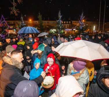 Na dworze wieczór. Mężczyzna z koszykiem wypełnionym opłatkiem przechodzi obok tłumu zgromadzonego na placu