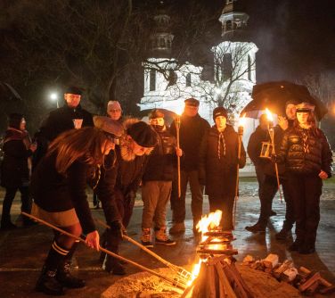 Na dworze wieczór. Na tle kościoła osoby stoją z pochodniami. Mężczyzna i kobieta zapalają od pochodni ognisko