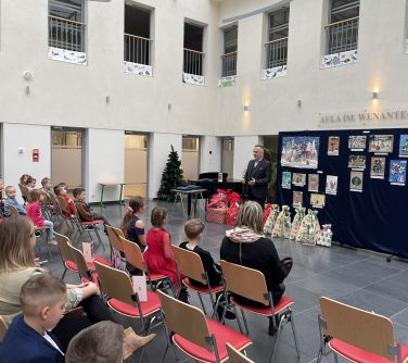 Aula. Mężczyzna na tle zwieszonych prac plastycznych oraz rozłozonych prezentów na podłodze przemawia do zgromadzonych osób...