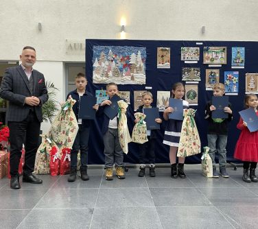 Aula. Mężczyzna oraz szóstka dzieci pozują do zdjęcia na tle zawieszonych prac plastycznych. Dzieci trzymają dyplomy i...