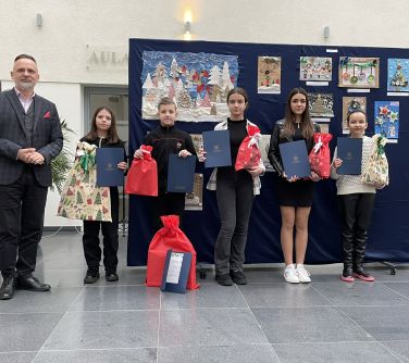 Aula. Mężczyzna oraz piątka dzieci pozują do zdjęcia na tle zawieszonych prac plastycznych dzieci. Dzieci trzymają dyplomy...