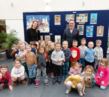 Aula. Grupa przedszkolaków, kobieta i mężczyzna pozują do zdjęcia na tle zawieszonych prac plastycznych