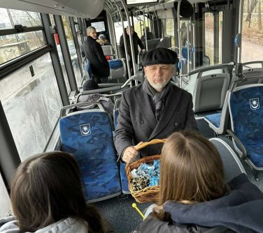 Wnętrze autobusu. Trzy osoby siedzą na fotelach. Mężczyzna w berecie z koszykiem wypełnionym gadżetami i cukierkami...