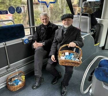Wnętrze autobusu. Mężczyzna w berecie z koszykiem na kolanach wypełnionym gadżetami i cukierkami oraz drugi uśmiechnięty...