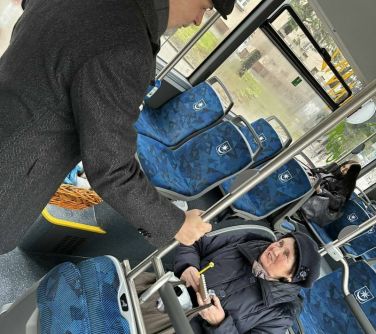 Wnętrze autobusu. Mężczyzna w berecie trzymając się za poręcz rozmawia z siedzącą kobietą