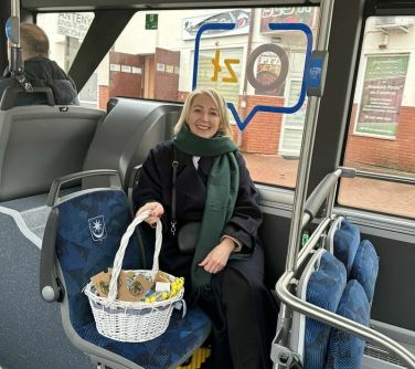 Wnętrzę autobusu. Uśmiechnięta kobieta siedzi na fotelu, trzyma za uchwyt koszyka wiklinowego wypełnionego gadżetami...
