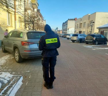 Na dworze w ciągu dnia, na dreptaku strażnik miejski tyłem, stoi za samochodem