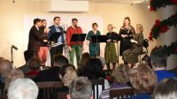 Aula. Na scenie osiem osób śpiewa do mikrofonów. Przed nimi pulpity. Przed sceną na krzesłach siedzą widzowie