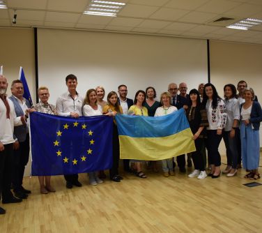 Pomieszczenie, aula. Grupa osób pozuje do zdjęcia. Z przodu trzymają flage Ukrainy i Unii Europejskiej