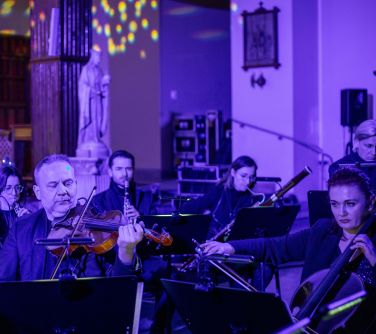 Wnętrze Kościoła. Orkiestra siedzi i gra na instrumentach. Przed nimi pulpity z nutami