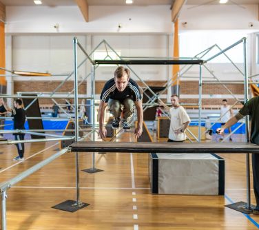 Hala sportowa. Rozstawione rusztowania do parkour. Nastolatek wskakuje na podest. Po prawej stronie stoi mężczyzna w czapce