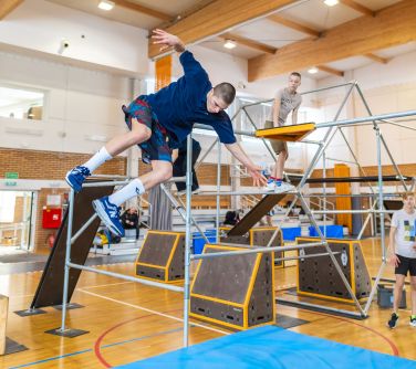 Hala sportowa. Rozstawione rusztowania do parkour. Pięcioro uczestników skacze po rurach