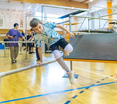 Hala sportowa. Rozstawione rusztowania do parkour. Chłopiec skacze nad rurą. W tle osoby