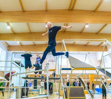 Hala sportowa. Rozstawione rusztowania do parkour. Mężczyzna skacze pomiędzy rusztowaniami. W tle po sali przechadzają się...