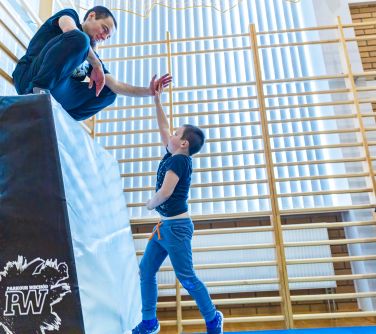 Hala sportowa. Rozstawione rusztowania do parkour. Chłopie przybija piątke z mężczyzną kucającym na wzniesieniu piankowym...