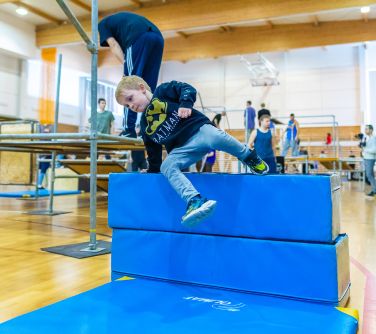 Hala sportowa. Rozstawione rusztowania do parkour. Chłopiec w dresie skacze przez wysoką piankową przeszkodę. W tle osoby