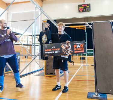 Hala sportowa. Rozstawione rusztowania do parkour. Uśmiechnięty nastolatek z kręconymi, krótkimi włosami jedną ręką trzyma...