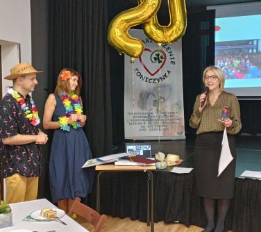 Aula. Na przodzie kobieta w czarnej spódnicy midi przemawia do mikrofonu. Po lewej stronie stoi usmiechnięta kobieta i...