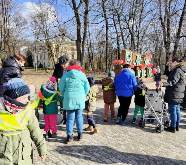 Na dworze. Grupa dorosłych wraz z dziećmi przechadza się chodnikiem. Wśród nich mężczyzna w kaszkiecie. W tle napis Mińsk...