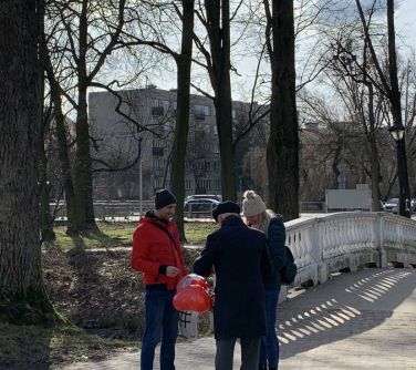 Na dworze. Na chodniku dwóch mężczyzn i kobieta. Mężczyzna odwrócony tyłem, trzyma koszyk udekorowany czerwonym balonem w...