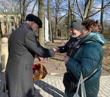 Na dworze. Mężczyzna w płaszczu i czapce wręcza upominek parze. W tle drzewa i budynek