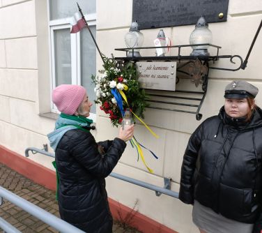 Na dworze, przy tablicy upamiętniającej. Harcerka stoi na posterunku. Kobieta w różowej czapce stawia zapalonego znicza pod...