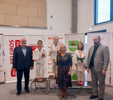 Hala sportowa. Na podium trzy dziewczynki w strojach judo, trzymają dyplomy. Przed podium stoi kobieta. Po obu stronach...