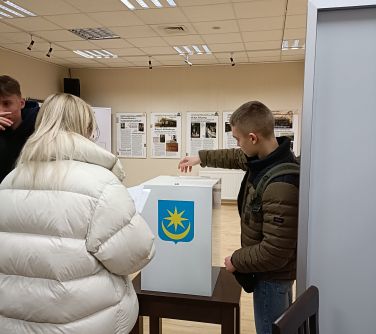 Pomieszczenie. Na małym ciemnobrązowym stoliku biała urna wyborcza z herbem Mińska Mazowieckiego. Nad nią nastolatek, który...