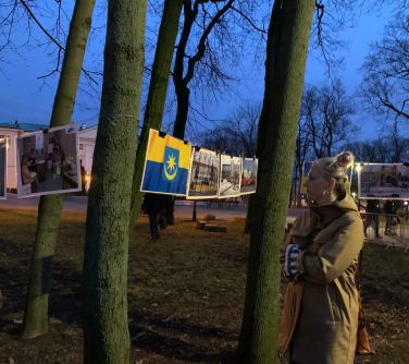Park pod wieczór. Pomiędzy drzewami na linkach zaczepione zdjęcia zniszczonej Ukrainy. Po prawej kobieta w jasnym płaszczu,...