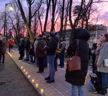 Alejka w parku wieczorową porą. Na niebie piękny zachód słońca, niebo różowe. Na alejce z kostki stoją osoby. Wzdłóż alejki...