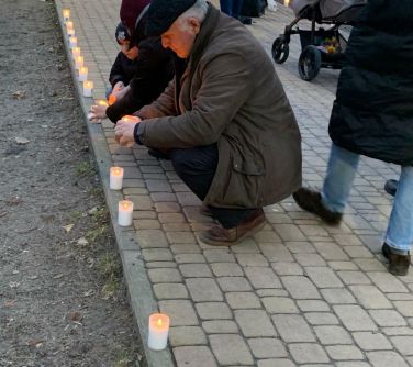 Alejka w parku. Kucający mężczyzna zapala kolejny znicz, który stworzy szpaler zapalonych zniczy. W tle przechadzający sie...