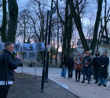 Park pod wieczór. Po lewej stojąc na ziemi przemawia do mikrofonu mężczyzna. Z jego lewego boku między drzewami rozwieszone...