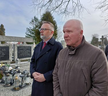 Cmentarz. Dwóch mężczyzn w zadumie. Mężczyzna po lewej w ciemnym płaszczu, okularach, z siwą brudką i włosami. Mężczyzna po...