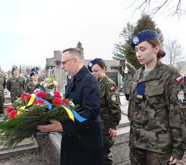 Cmentarz. Mężczyzna w okularach i ciemnym płaszczu trzyma wiązankę biało-czerwonych kwiatów z niebiesko-żółtą szarfą. Po...
