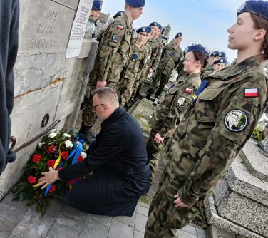 Cmentarz. Mężczyzna w ciemnym płaszczu kładzie wiązankę z biało-czerwonych kwiatów z szarfą niebieską- żółtą pod pomnikiem...