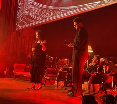 Scena podświetlona na czerwono. Kobieta z ciemnymi włosami, w czarnej sukni wskazuje dłonią na stojącego obok mężczyznę w...