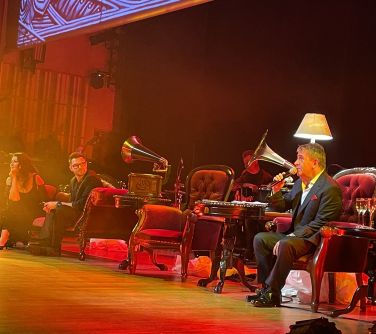 Scena. Dwa fotele i sofa. Pomiędzy dwa stare gramofony i zapalona stojąca lampa. Na jednym z foteli siedzi i śpiewa...