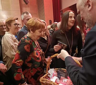 Sala koncertowa. Po koncercie mężczyzna trzyma koszyk wiklinowy, wręcza wychodzącej widowni upominki