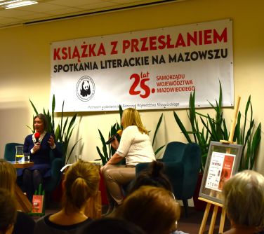 Sala konferencyjna. Na dwóch fotelach siedzą kobiety. Po lewej kobieta przemawia do mikrofonu. Przed nimi stolik. W tle na...