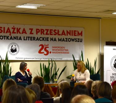 Sala konferencyjna. Na dwóch fotelach siedzą kobiety. Po lewej stronie kobieta przemawia do mikrofonu. Przed nimi stolik na...