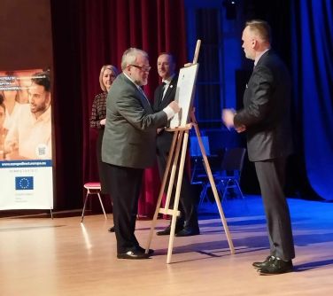 Scena. Na drewnianej sztaludze duży obrazek z tytułem Deklaracja. Mężczyzna podpisuje dokument. Po bokach stoją trzy osoby...