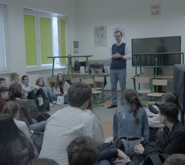 Sala lekcyjna. Zsunięte ławki i krzesła. Na przodzie stoi mężczyzna i opowiada. Na podłodze siedzą nastolatkowie. Po prawej...