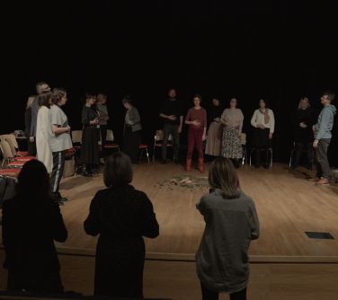 Aula, scena. Na scenie w półkolu stoją osoby, za nimi krzesła. Po środku wysypane gałązki, szyszki, płatki. Przed sceną...