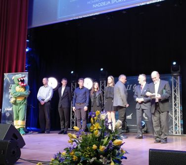 Na scenie stoi osiem osób. Po lewej dziewiąta osoba przebrana za smoka. Na przodzie dwie niskie kolumny, między nimi bukiet...