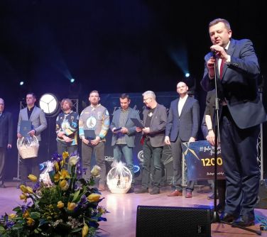 Na scenie stoi siedem osób. Ósma osoba, mężczyzna przemawia do mikrofonu. Na przodzie bukiet wiosennych kwiatów