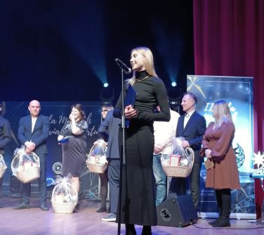 Scena. Kobieta w czarnej sukience przemawia do mikrofonu. W tle stoją pozostałe osoby z koszami prezentowymi